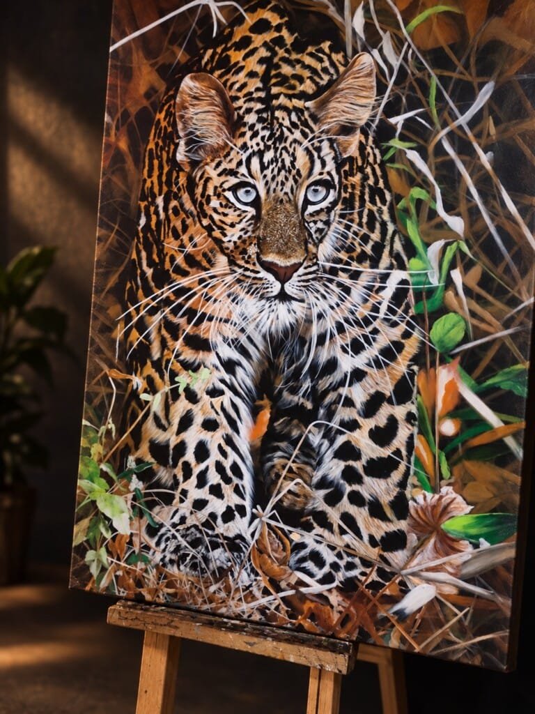 THE LONER by Sumana Burman, an original wildlife realism painting of a leopard emerging through dense forest foliage, displayed on a wooden easel.