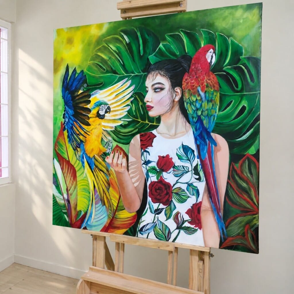 WILDERNESS by Sumana Burman, an original contemporary realism painting of a woman with colorful parrots and tropical foliage, displayed on an easel.